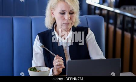 Older Woman Analyzing Data While Eating. Retired Businesswoman Multitasks Using Laptop During Lunch Break. Senior Female Entrepreneur Focuses On Stock Photo