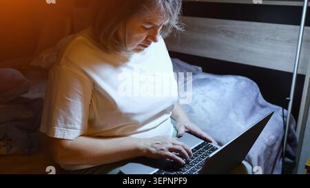 Mature woman in pajamas typing on a laptop in bed at night, bathed in screen glow, working or browsing online late at home, comfortable and connected Stock Photo