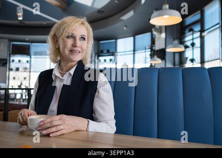 Senior Businesswoman Enjoying Quiet Cafe Moment Seated In Blue Booth With Natural Light, Smiling Softly While Holding Cup And Resting Hands On Table Stock Photo