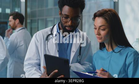 Two multiracial doctors Caucasian woman intern with African man therapist talking using digital tablet look in papers result discuss medicine health Stock Photo