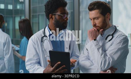 Two multiethnic doctors men pensive discussing medicine test results looking in digital tablet in clinic. African American therapist and Caucasian Stock Photo