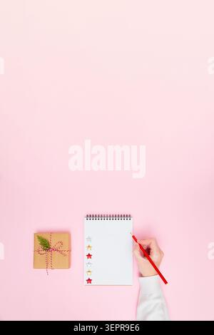 Female hand writing on blank notepad with gift box on pastel pink background, festive flat lay vertical banner with copy space for holiday planning, goals, to-do list or wish list. Stock Photo