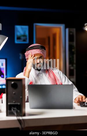 Tired male entrepreneur in Arabic attire browses the internet on a laptop, frustrated, he touches his forehead; despite feeling sick, Middle Eastern man remains committed to his work in modern office. Stock Photo