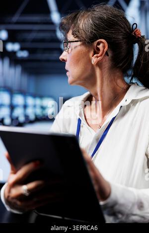 Old computer scientist updating data center rackmounts, writing intricate binary code scripts on tablet, using programming to fortify cybersecurity in technically advanced supercomputer hub. Stock Photo