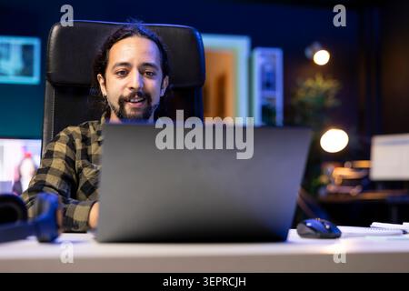 Male individual in his home environment, smiling while checking emails on a laptop, portrays the happiness and ease that technology brings to his daily life; cheerful guy browsing the internet. Stock Photo