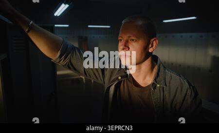 Cinematic Close Up of Focused Male Athlete Leaning Hand on Metal Locker, Looking Thoughtfully Into Window With Warm Morning Light on Face Inside Sports Club. Concept of Authentic Lifestyle Moment. Stock Photo