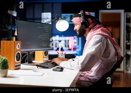 Middle Eastern software programmer typing code on a laptop showcasing the essence of software programming and development while arab man with wireless headphones engrossed in running algorithms. Stock Photo