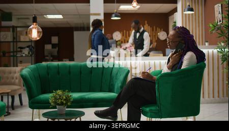Worker managing bookings by phone, answering calls to register to clients for room reservation and writing personal information on clipboard. Hotel staff taking notes using telephone line. Stock Photo