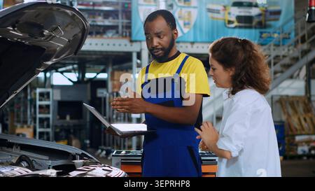 Experienced mechanic using laptop to show woman damaged components found in her car. Stock Photo
