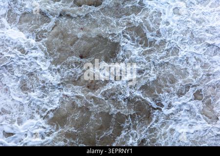 White foam forms on the surface of the water as waves crash and recede. The water appears choppy with a mixture of colors and textures in the current. Stock Photo