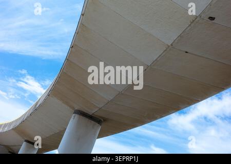 The scene shows a curved concrete structure viewed from below under a blue sky filled with soft white clouds. This structure is part of an urban lands Stock Photo