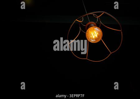 A light bulb hangs from a metal fixture in a dark space. The warm glow of the bulb stands out against the black background, illuminating the area belo Stock Photo