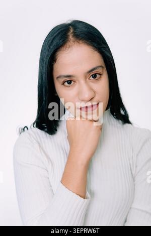 Young Southeast Asian brunette woman over isolated white background resting chin on her fist and looking forward with a serious or calm expression. Stock Photo