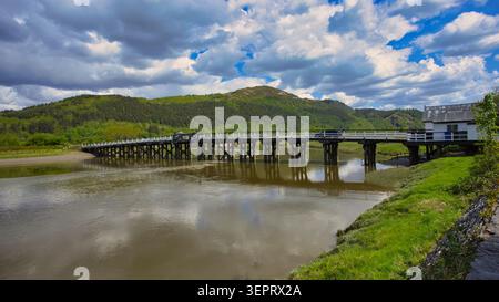 Scenic wooden Penmaenpool Toll Bridge over the Mawddach Estuary, in North Wales set against green hills under a cloudy sky. Stock Photo