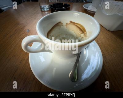 Empty white coffee cup on a saucer on a wooden cafe table, with espresso residue, a spoon, glass, ashtray and sugar holder visible in the background o Stock Photo