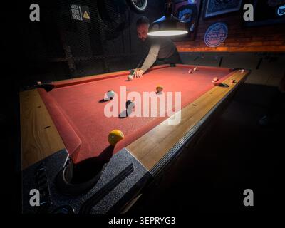 BELGRADE, SERBIA - MARCH 28, 2026: man playing pool on a red felt pool table in a dimly lit bar, with billiard balls, cue stick and pub interior light Stock Photo