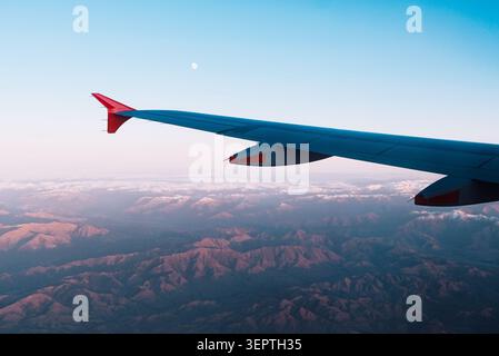 South Island, New Zealand: Aerial view from an airplane window seat of the wing over rugged mountain terrain under a blue twilight sky with the moon. Stock Photo