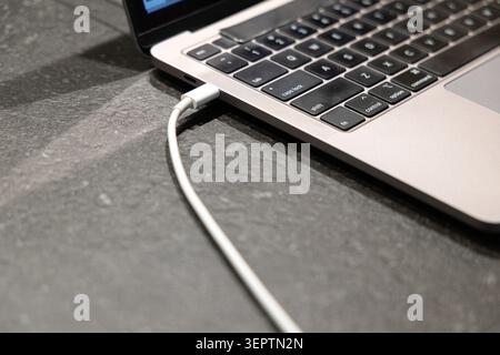 Close-up of a silver laptop partially open on a dark textured stone surface, with a white USB-C charging cable plugged in, representing modern telework and remote work lifestyle. Stock Photo