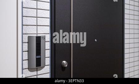 A modern Japanese rental apartment entrance door numbered 704 with a wall-mounted intercom and speaker panel stands against a white textured tile wall. A typical residential entrance documented during hikkoshi (Japanese moving day) in an urban condominium building. Stock Photo