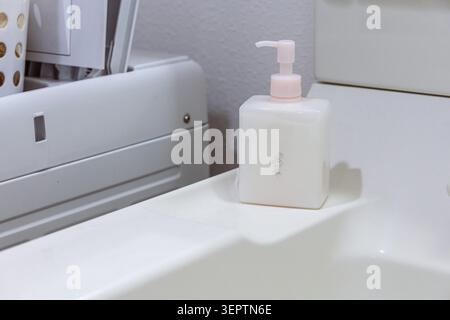 A white square pump soap dispenser with a delicate rose motif sits on a clean bathroom vanity ledge, with white bathroom accessories softly blurred in the background. Stock Photo