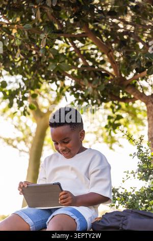 African American boy child sitting under leafy tree at park holding tablet, backpack on right Stock Photo