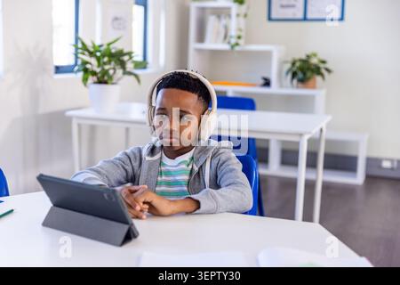 African American boy sitting at table using tablet in classroom wearing hoodie and over-ear headset Stock Photo