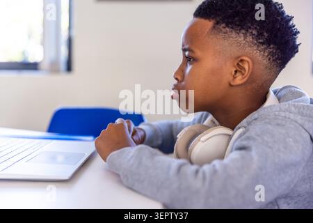 African American boy child sitting at school using silver laptop on white table, wearing headphones Stock Photo