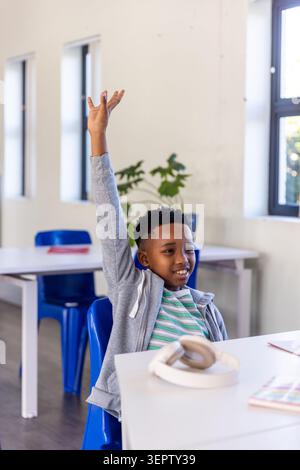 African American child sitting in class raising right hand at desk with headphones and notebook Stock Photo