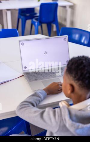 School-age boy sitting at classroom table using silver laptop showing center crosshair, copy space Stock Photo