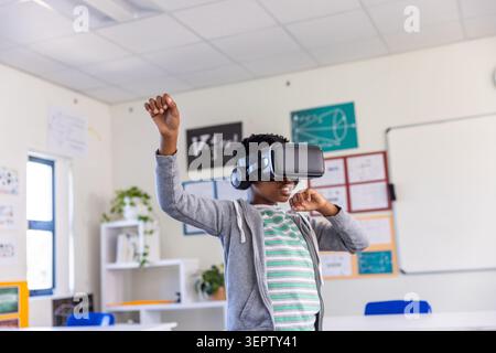 African American boy standing in classroom, wearing VR headset with headphones, hoodie, raising arm Stock Photo