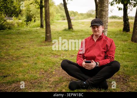 Man sitting on grass, leaning against a tree, checking his phone in nature Stock Photo