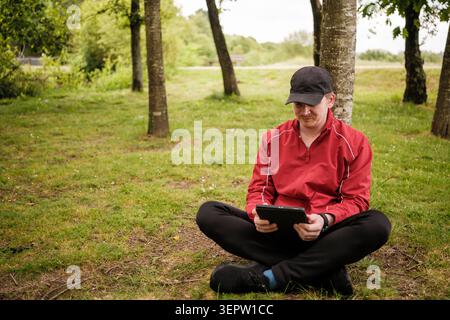 Man sitting on grass, leaning against tree, using a tablet for leisure outdoors Stock Photo