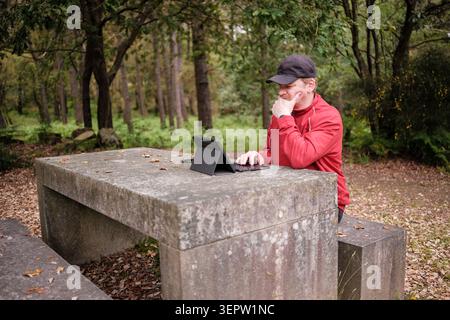 Man using a digital tablet with keyboard at a stone picnic table in a forest Stock Photo