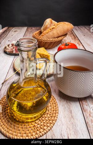 Glass olive oil cruet and white enamel pitcher beside bread basket, ham, tortilla chunks and tomato on whitewashed wood. Stock Photo