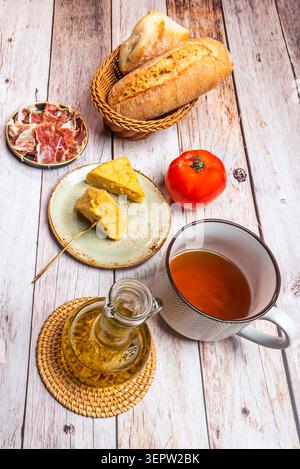 Glass olive oil cruet and white enamel pitcher beside bread basket, ham, tortilla chunks and tomato Stock Photo