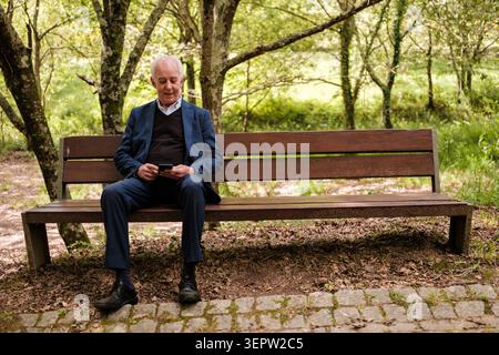 Senior man sitting on a wooden bench in a park, engaging with his smartphone Stock Photo