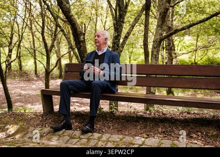 Senior man sitting on a park bench using a smartphone while looking away Stock Photo