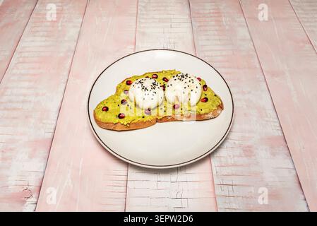 Smashed avocado toast topped with two burrata balls, sesame seeds and pomegranate sits on a white plate over pink wood. Stock Photo