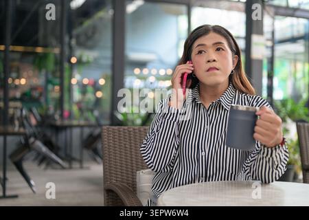 Thai Isan transgender businesswoman in 40s talking on phone at cafe veranda. Horizontal medium shot portrait of a mature non-binary Southeast Asian tr Stock Photo