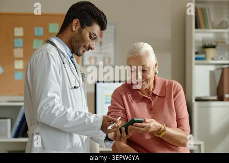 Senior Woman Visiting Doctor and Using Smartphone in Clinic Stock Photo