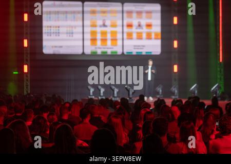 Conference speaker presents slides to large seated audience on stage, moody red lighting and blurred projection screen create focused atmosphere. Stock Photo