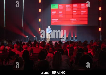 Business conference speaker presents on stage to large attentive audience, dramatic red lighting and projection screen. Stock Photo