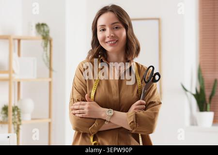 Smiling young female seamstress with measuring tape and scissors, fashion design and tailoring business, this professional woman embodies creativity a Stock Photo