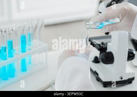 Scientist pouring blue liquid from test tube into another test tube in laboratory with microscope and rack Stock Photo