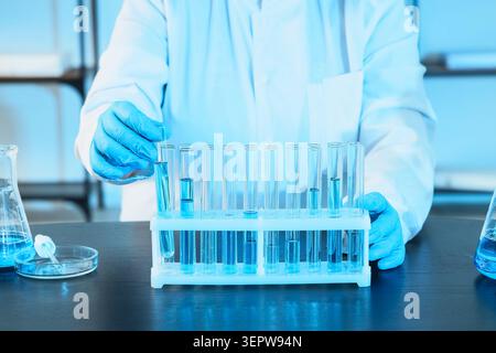 Scientist in lab coat and blue gloves handling test tube with blue liquid in research laboratory, the laboratory setting includes a rack of test tubes Stock Photo