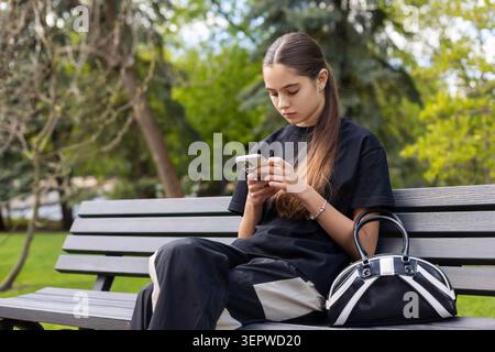 Young woman using smartphone on park bench. Young woman sits on a wooden park bench using a smartphone, with a black handbag beside her and soft green Stock Photo