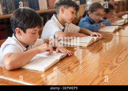 Group of elementary school students sitting at a wooden table and reading books in a library. Stock Photo
