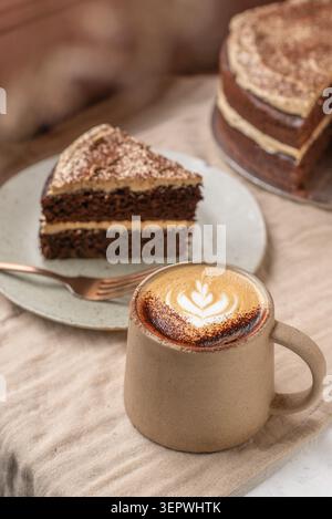 A slice of double-layer chocolate cake with cream frosting next to a hot cappuccino with leaf latte art, with the main cake in the background. Stock Photo