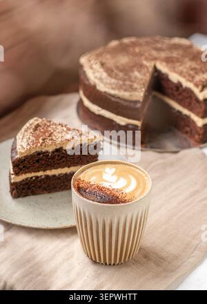 A slice of double-layer chocolate cake with cream frosting next to a hot cappuccino with leaf latte art, with the main cake in the background. Stock Photo