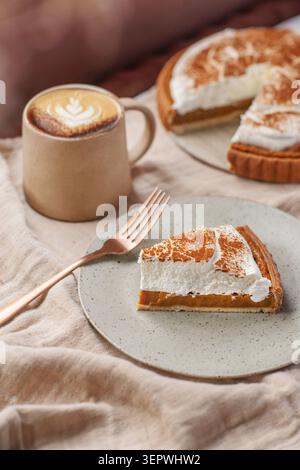 A slice of pumpkin pie topped with whipped cream and cinnamon next to a hot cappuccino with latte art, with the whole pie in the background. Stock Photo